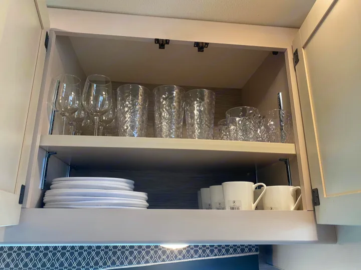 Inside an RV rental cabinet: a shelf of clear glasses above white plates and mugs, providing essential kitchenware.. Thor A.C.E 2020