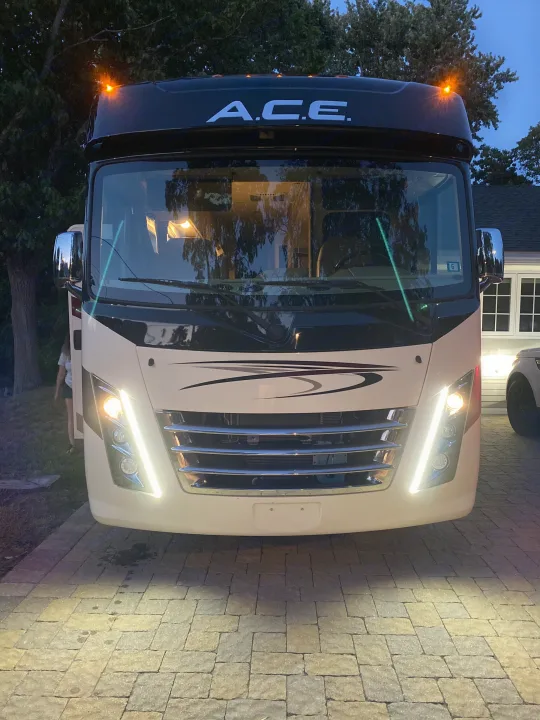 Front view of a cream-colored A.C.E. motorhome, possibly an RV rental, parked on a paver driveway at dusk with its bright lights on.. Thor A.C.E 2020