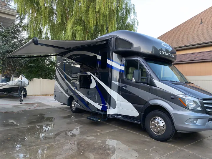 A large gray Citation RV with an extended awning is parked on a wet driveway next to a boat, perfect for an rv rental.. Thor Motor Coach Citation 2018
