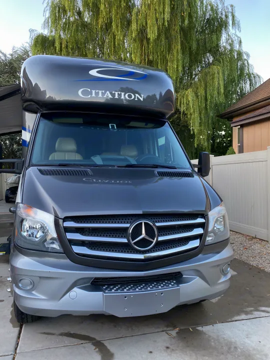 A sleek gray Mercedes-Benz Citation RV, excellent for rv rental, seen from the front, parked next to a house and willow tree.. Thor Motor Coach Citation 2018