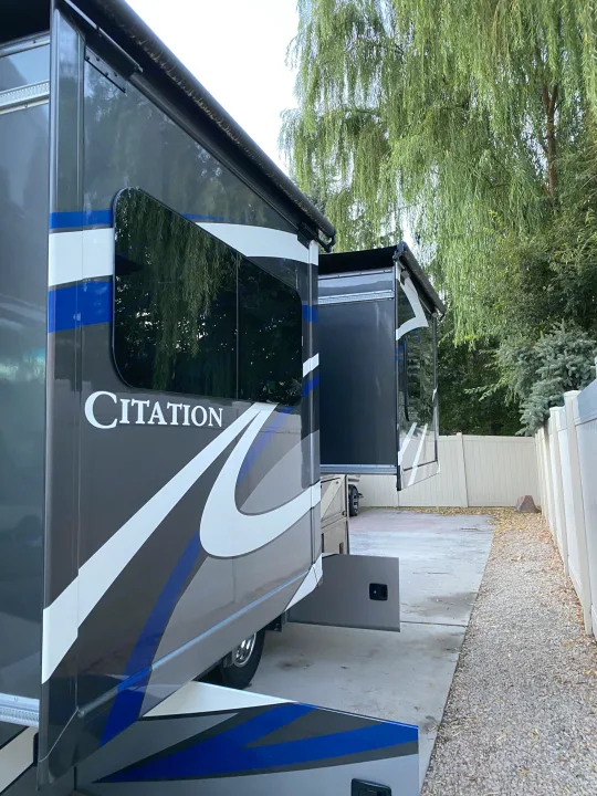Grey Citation RV with blue stripes & extended slide-outs, ready for your next rv rental adventure under a willow tree.. Thor Motor Coach Citation 2018