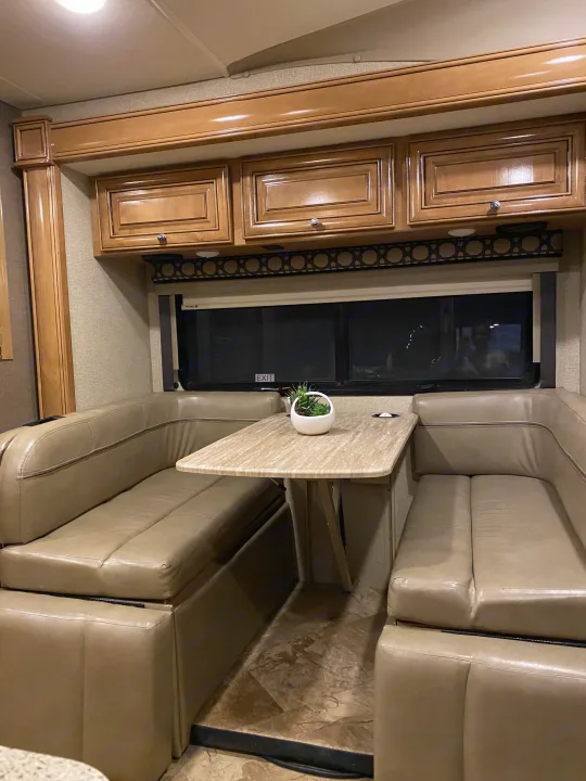 This RV rental dinette features U-shaped beige leather seating, a light table, wood cabinets, and a window.. Thor Motor Coach Citation 2018