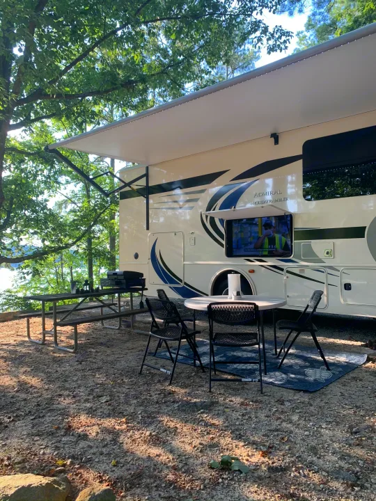 RV rental set up by a lake, featuring an outdoor TV, extended awning, grill, and picnic table for camping.. Holiday Rambler Admiral 2021