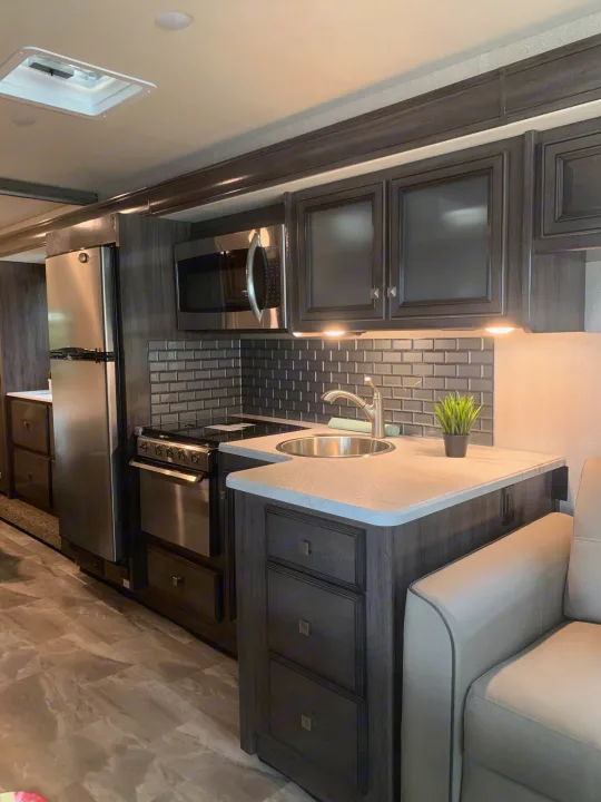 Sleek RV rental kitchen & living area with stainless steel appliances, dark cabinetry, and light sofa.. Holiday Rambler Admiral 2021