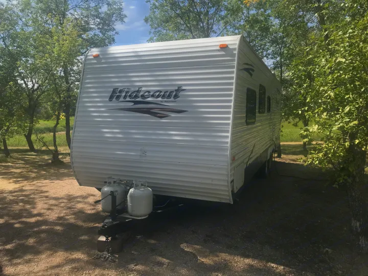 White Hideout travel trailer (RV rental) in a shady campsite, with propane tanks visible at the front, surrounded by trees.. Keystone Hideout 2009