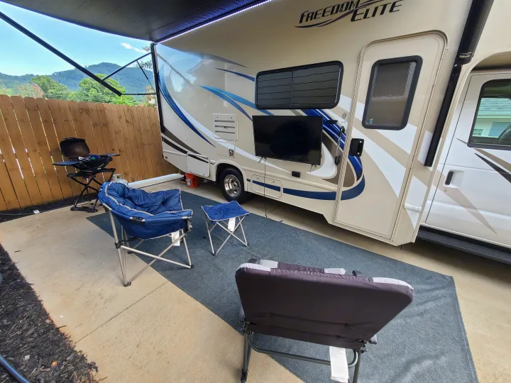 An RV rental set up on a patio with an extended awning, outdoor TV, grill, and chairs, offering mountain views.. Thor Freedom Elite 2019