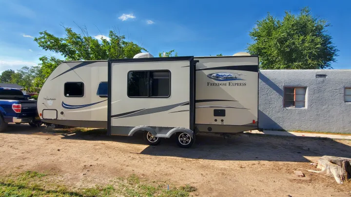 A beige Coachmen travel trailer hooked to a blue pickup truck on a dirt lot, ready for an RV rental trip under a blue sky.. Coachmen Freedom Express 2015