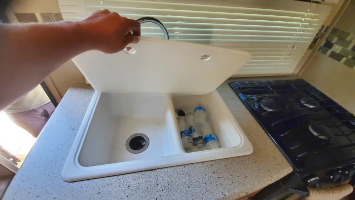 Person lifts sink cover in an RV rental kitchen, revealing double-basin sink with bottles and stove.. Coachmen Freedom Express 2015