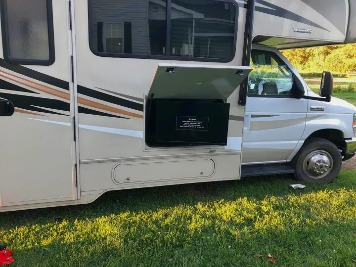 White and tan RV rental parked on grass, showing an open side compartment with a TV screen for outdoor viewing.. Winnebago Minnie Winnie 2017