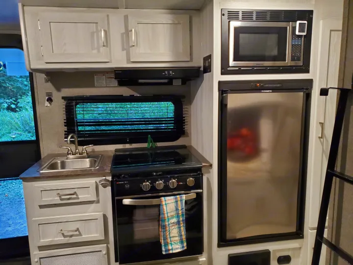 An rv rental kitchen with a sink, stove, microwave, and refrigerator, all framed by light wood-grain cabinets and a window view.. Forest River Rockwood Geo Pro 2021
