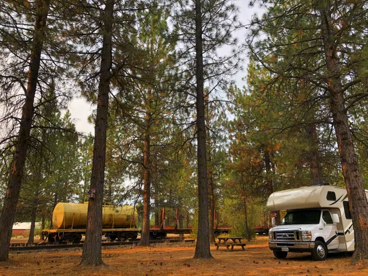 Scenic **RV rental** parked among tall pines near a historic train track with old rail cars and a picnic table. Perfect for a quiet getaway.. Other Four Winds 2018