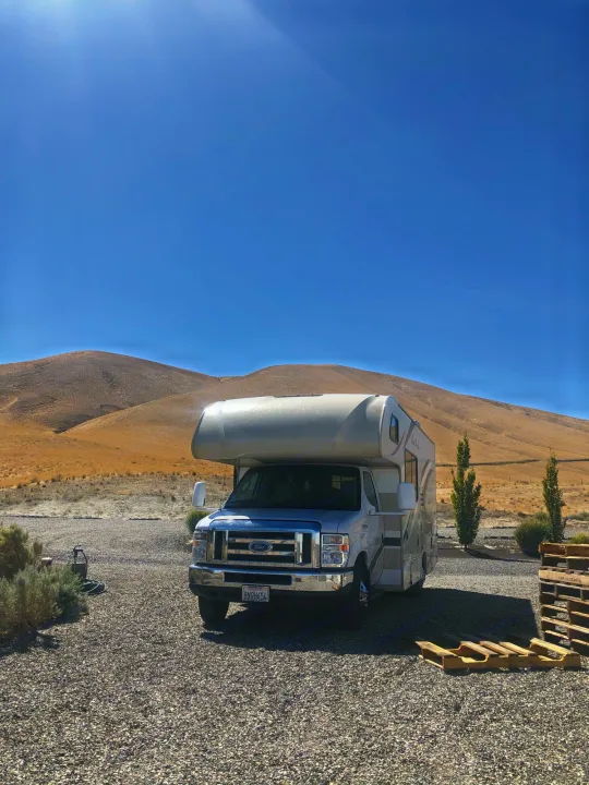 This RV rental is parked on a gravel lot, backed by dry, rolling hills under a clear blue sky, ideal for your next trip.. Other Four Winds 2018
