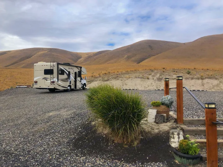 An RV rental is parked on a gravel lot, framed by dry, rolling hills. A green bush and steps with solar lights are in the foreground.. Other Four Winds 2018