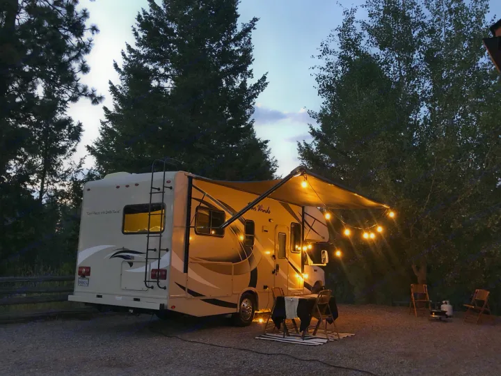 Cozy RV rental campsite at dusk: a motorhome with illuminated awning, outdoor dining, and lounge area among trees.. Other Four Winds 2018