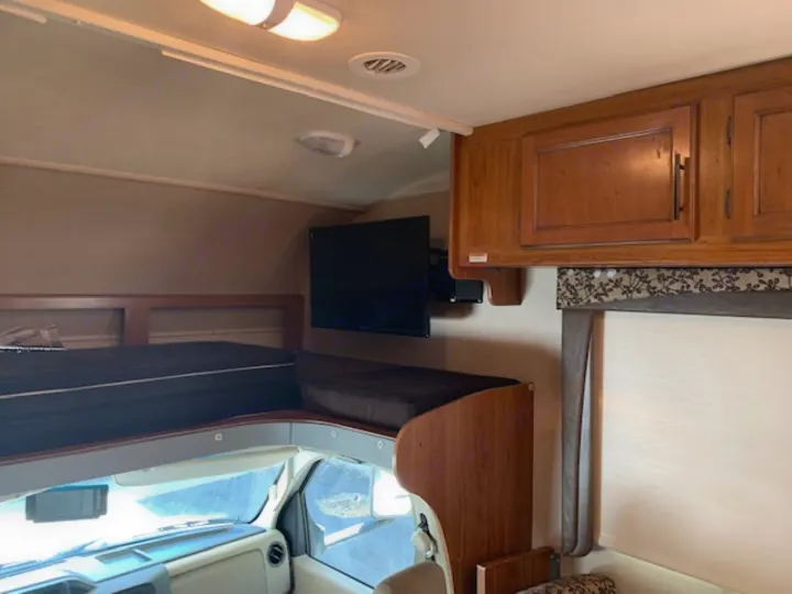 RV rental bunk area with TV, overhead cabinets, and window blind, offering a compact sleeping and living space above the cab.. Jayco Redhawk 31X 2017