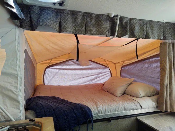 Inside a cozy RV rental: a plush bed with soft pillows & blanket in an expandable camper sleeping area.. Forest River Rockwood Freedom 2021