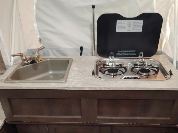RV rental pop-up camper kitchen featuring a stainless steel sink, faucet, two-burner stove, and light countertop.. Forest River Rockwood Freedom 2021