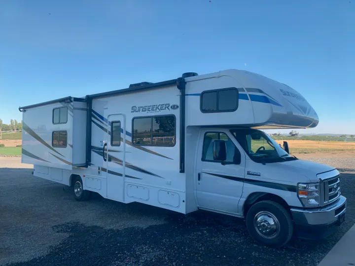 This white Forest River Sunseeker Class C RV rental, with an extended slide-out, is parked on gravel under a clear blue sky.. Forest River Sunseeker 2021
