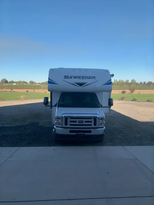 Front view of a white Sunseeker RV on a gravel driveway, with fields and clear sky in the background. Ideal for your next RV rental adventure.. Forest River Sunseeker 2021