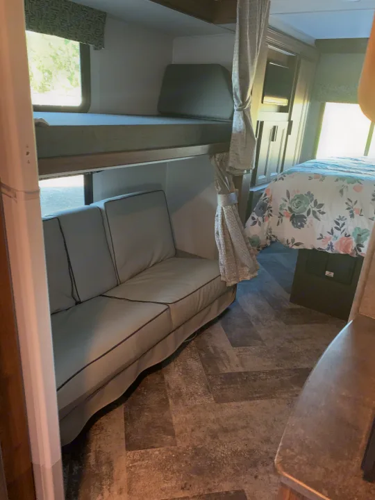 Interior of an RV rental: bunk beds, gray sofa, and master bed with floral comforter visible, all on wood-look flooring.. Forest River Sunseeker 2021