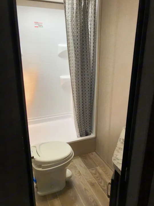 An RV rental bathroom showing a white toilet, shower stall with a gray patterned curtain, and light wood-look floor.. Jayco Jay Flight 2021
