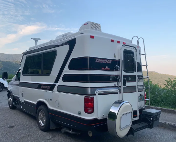 Rear view of a white Chinook rv rental camper van with roof AC, ladder, & spare tire, parked in a scenic spot.. Chinook Concourse 2001