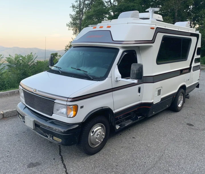 White Chinook RV rental, a compact motorhome, parked on asphalt with scenic mountains and trees in the background.. Chinook Concourse 2001