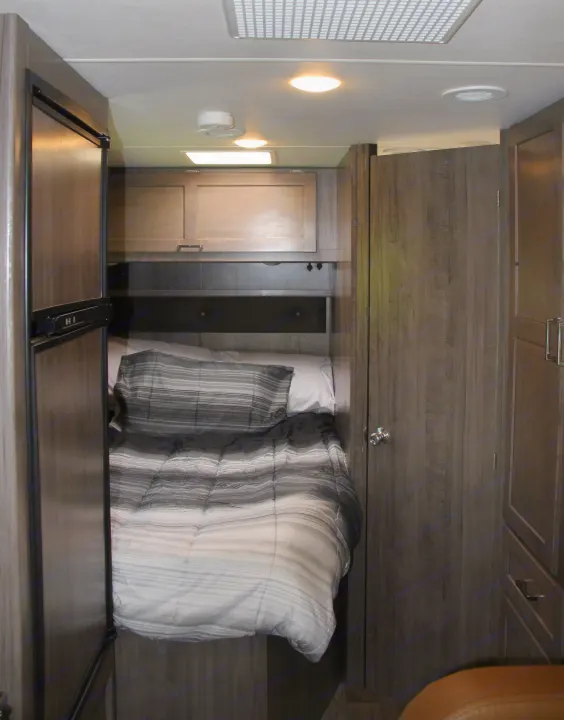 Compact RV rental bedroom featuring a striped bed, refrigerator, overhead cabinets, and ceiling fixtures.. Winnebago Minnie Winnie-Hook 2019