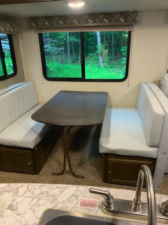 RV rental dinette with dark table, white benches, and a forest view. Kitchen sink has a note about flushing the fresh water system.. Keystone Bullet 2018