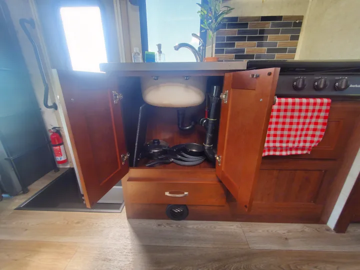 RV rental kitchen interior: open sink cabinet reveals pots and pans. Stove with red-checkered towel, counter, and fire extinguisher.. Forest River Sunseeker 2018