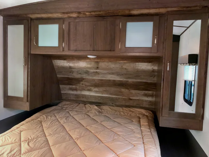 Relax in this RV rental bedroom with a rustic headboard, frosted/mirrored cabinets, and a soft comforter.. Keystone Passport 2020
