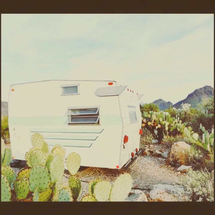 A white vintage travel trailer, a perfect RV rental, stands among prickly pear cacti in a peaceful desert with mountains.. Shasta Compact 1971