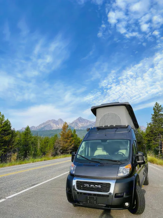 Experience nature with an RV rental! Dark camper van, pop-top up, on a scenic road with mountains & blue sky.. Thor Tellaro 2020