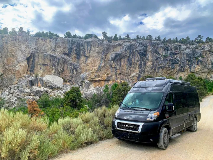 Black Ram camper van, perfect as an rv rental, parked on a dirt road beside a towering rock cliff. Ideal for off-road adventures.. Thor Tellaro 2020
