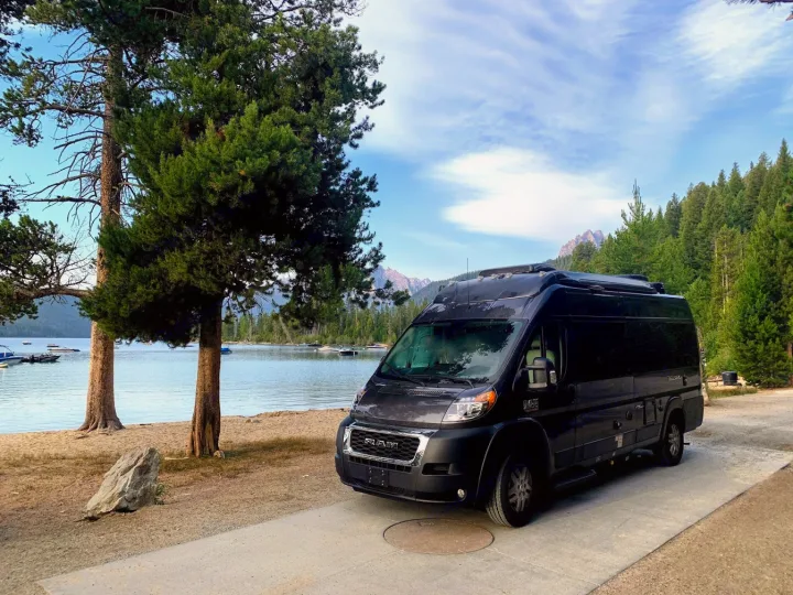 This black RV rental is parked by a tranquil lake with boats, flanked by tall trees and mountains. Ideal for scenic adventures.. Thor Tellaro 2020