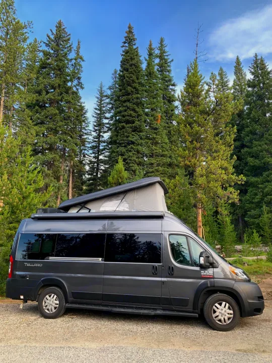 Gray pop-top camper van for rv rental, parked among tall trees. Perfect for camping in nature.. Thor Tellaro 2020