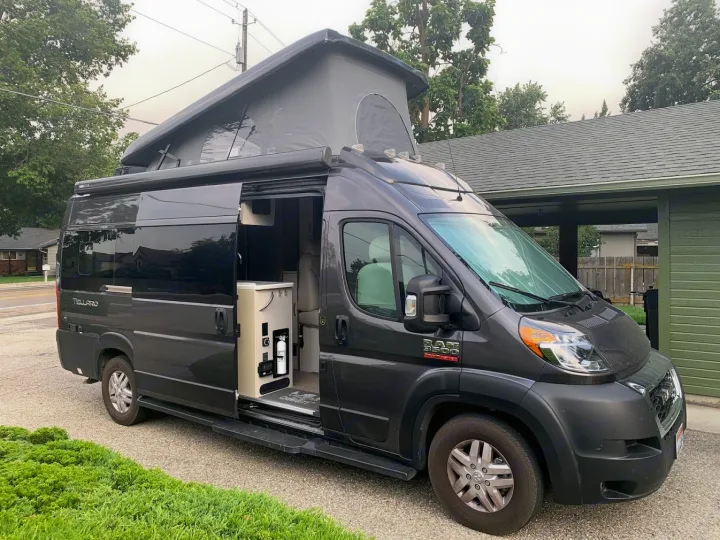 Dark gray RAM ProMaster pop-top RV rental with side door open, showcasing its outdoor kitchen unit.. Thor Tellaro 2020