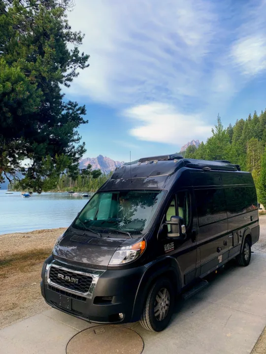 Scenic RV rental adventure! Black camper van parked by a serene lake with mountains & trees under a blue sky.. Thor Tellaro 2020