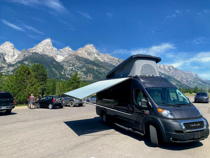 Dark gray RV rental with pop-top & awning deployed, parked in a lot with majestic Teton mountains & clear sky. Ready for adventure.. Thor Tellaro 2020