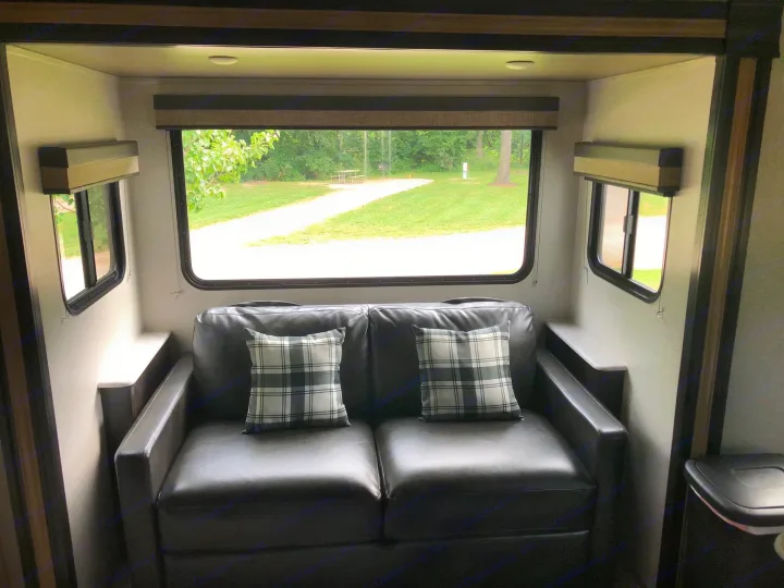 Inside a spacious RV rental, a cozy sofa with plaid pillows faces a large window overlooking a peaceful campsite.. ForestRiver Surveyor 2020