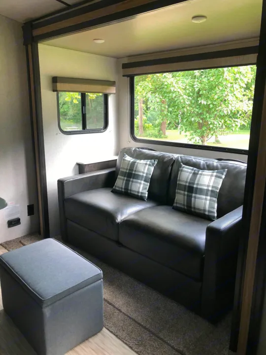 Cozy RV rental living space featuring a dark couch, plaid pillows, a storage ottoman, and windows with scenic views.. ForestRiver Surveyor 2020