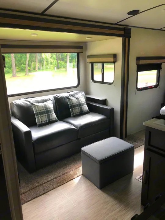 Cozy RV rental interior with a dark grey sofa, plaid pillows, and an ottoman, offering a view of nature outside.. ForestRiver Surveyor 2020