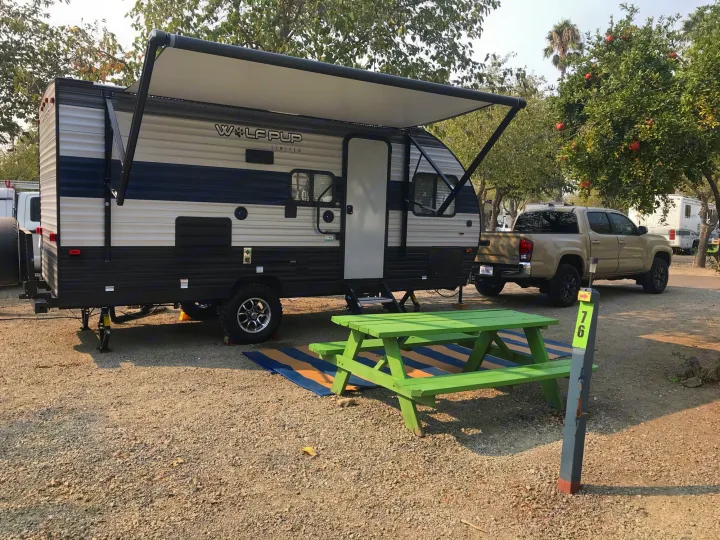 RV rental setup with a travel trailer, extended awning, green picnic table, and truck at a gravel campsite (site 76).. Forest River Wolf Pup 2021
