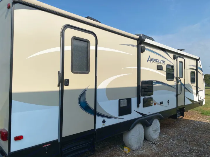 This beige Aerolite travel trailer, perfect for an rv rental, features stylish blue graphics and is parked on gravel.. Dutchmen Aerolite 2017