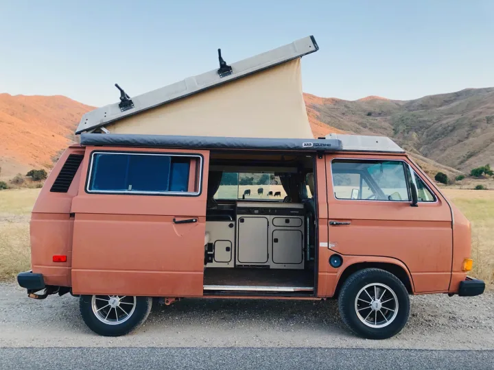 Scenic RV rental: Orange pop-top camper van with open door showing Westfalia kitchen. Mountains and cows in the background.. Volkswagen Westfalia 1982