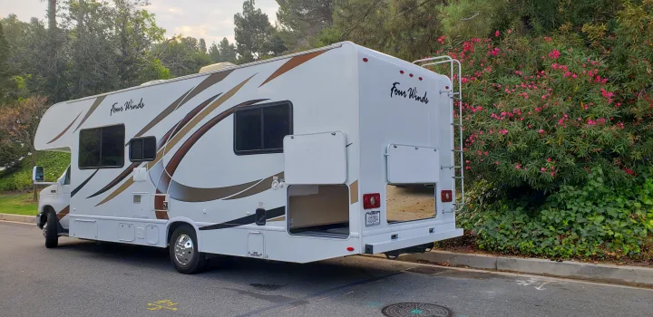 This white Four Winds RV rental, with brown graphics, is parked on a street near green bushes and pink flowers, storage open.. Thor 28A 2019