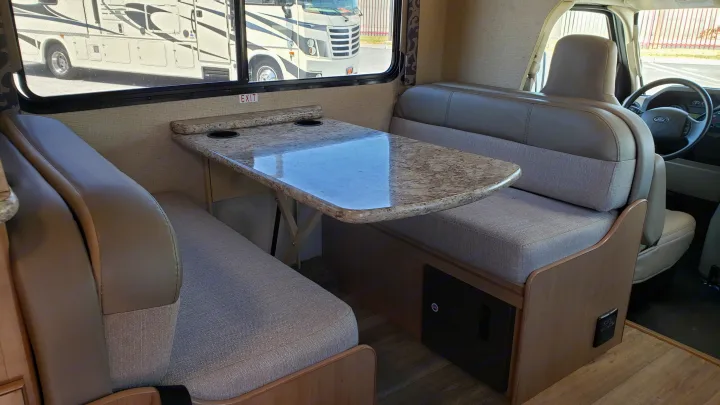 An rv rental interior featuring a dinette with two upholstered benches and a table; the driver's seat is visible in front.. Thor 28A 2019