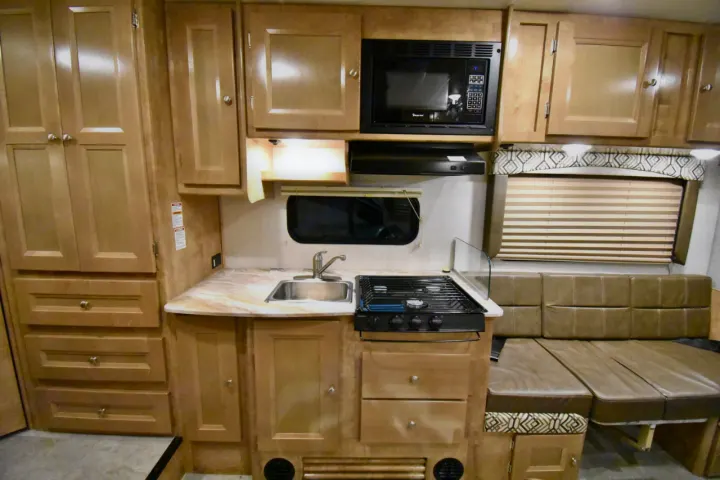 RV rental interior features a compact kitchen with stove, microwave, sink, and ample wood cabinets. Includes a cozy dinette.. Leprechaun 230RB 2020