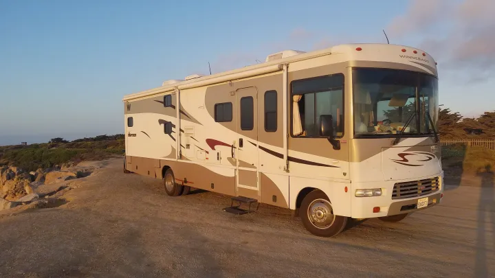A beige and white Winnebago Sightseer RV rental is parked on a dirt path next to a rocky coast under a clear sky.. Winnebago Sightseer 2007