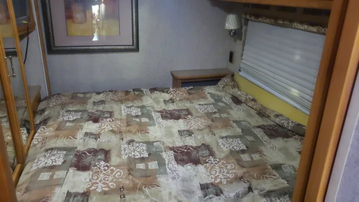 RV rental bedroom interior: Features a large bed with patterned comforter, nightstand, wall lamp, and window with pleated blinds.. Winnebago Sightseer 2007